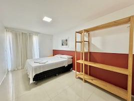 Una cama individual con una colcha blanca, una estantería de madera, un cuadro en la pared y una ventana con cortinas.