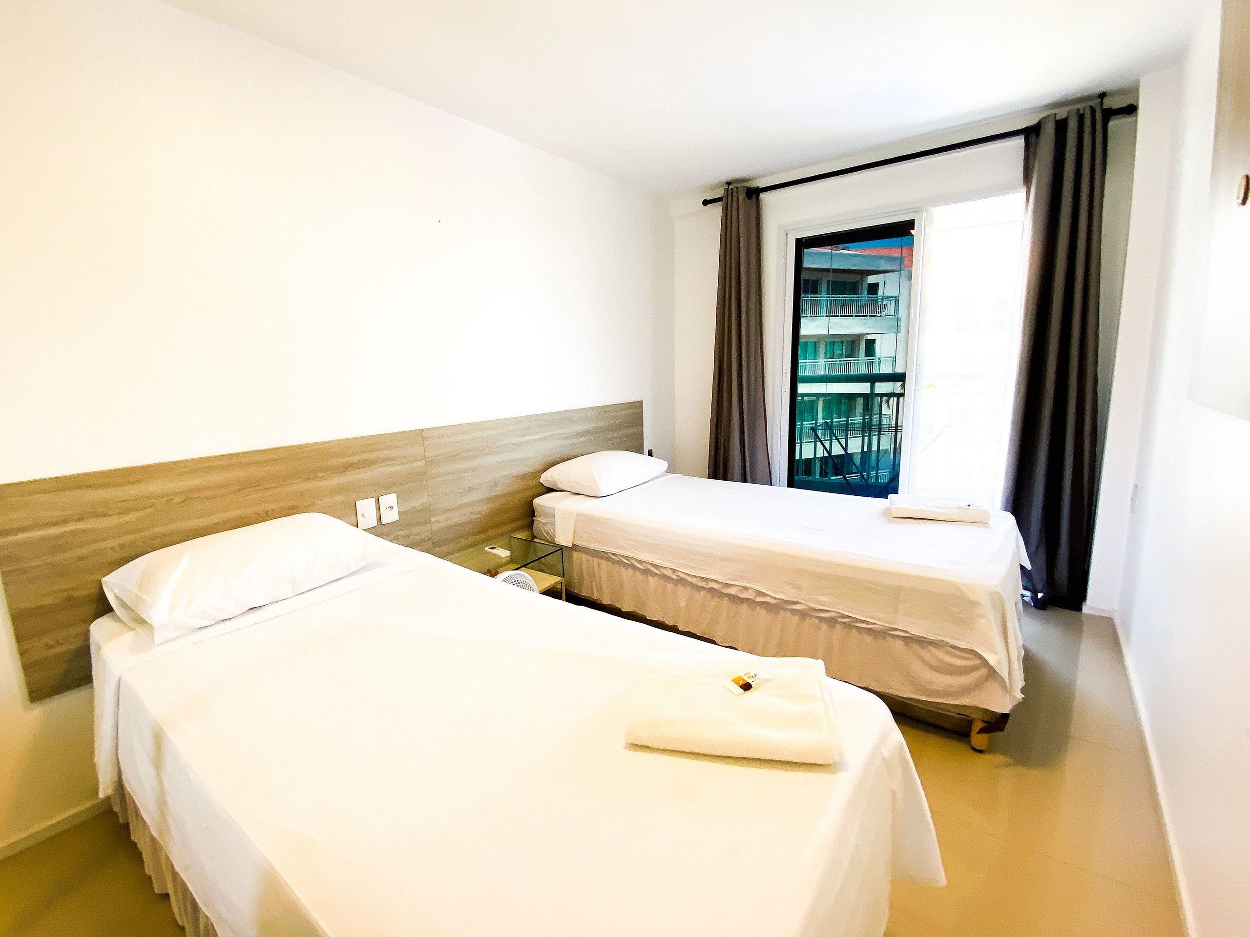 A hotel room with two single beds, a small table, a window with curtains, and a view of a building outside.