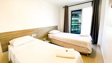 A hotel room with two single beds, a small table, a window with curtains, and a view of a building outside.