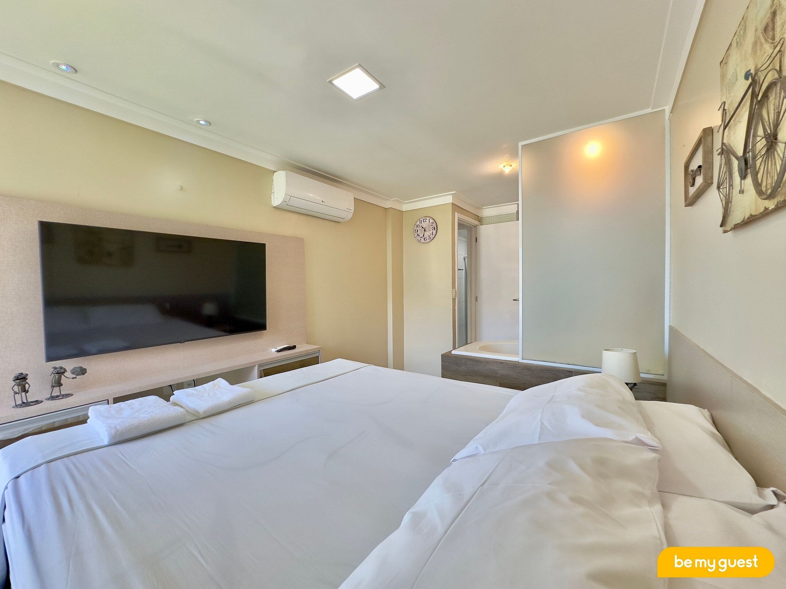 Habitación de hotel con una cama grande, un televisor de pantalla plana en la pared, zona de ducha y un reloj en la pared.