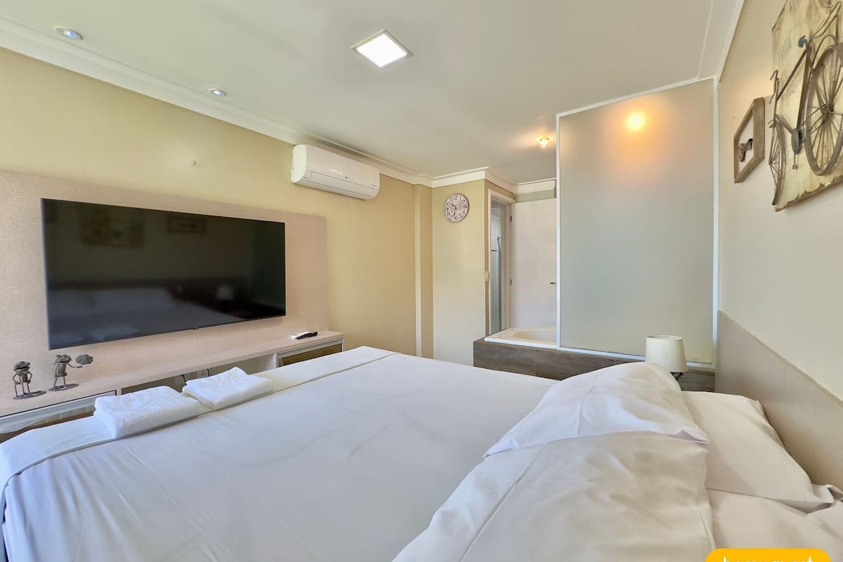 A hotel room with a large bed, a flat-screen TV mounted on the wall, a shower area, and a clock on the wall.