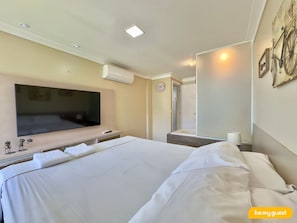 A hotel room with a large bed, a flat-screen TV mounted on the wall, a shower area, and a clock on the wall. - Solarium Residence Resort (Aquiraz)