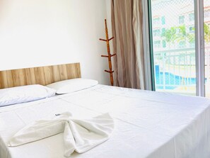 Habitación de hotel con cabecera de madera, cama con ropa blanca, una toalla sobre la cama y vista a una piscina a través de una puerta de vidrio.