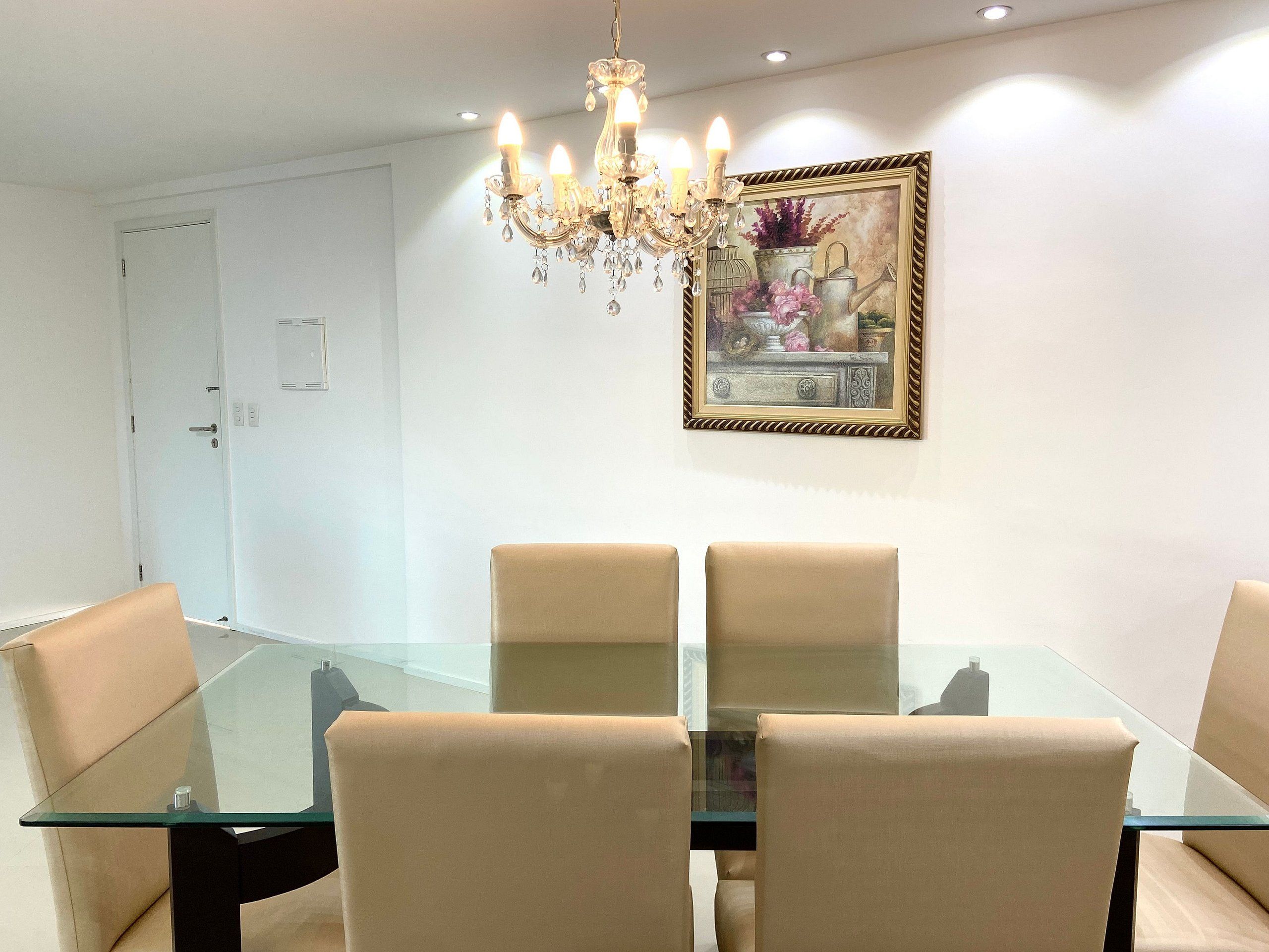 A dining area with a glass table and beige chairs, a chandelier, and a framed painting on the wall.