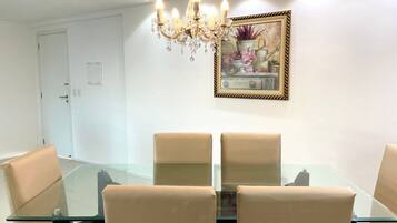 A dining area with a glass table and beige chairs, a chandelier, and a framed painting on the wall.