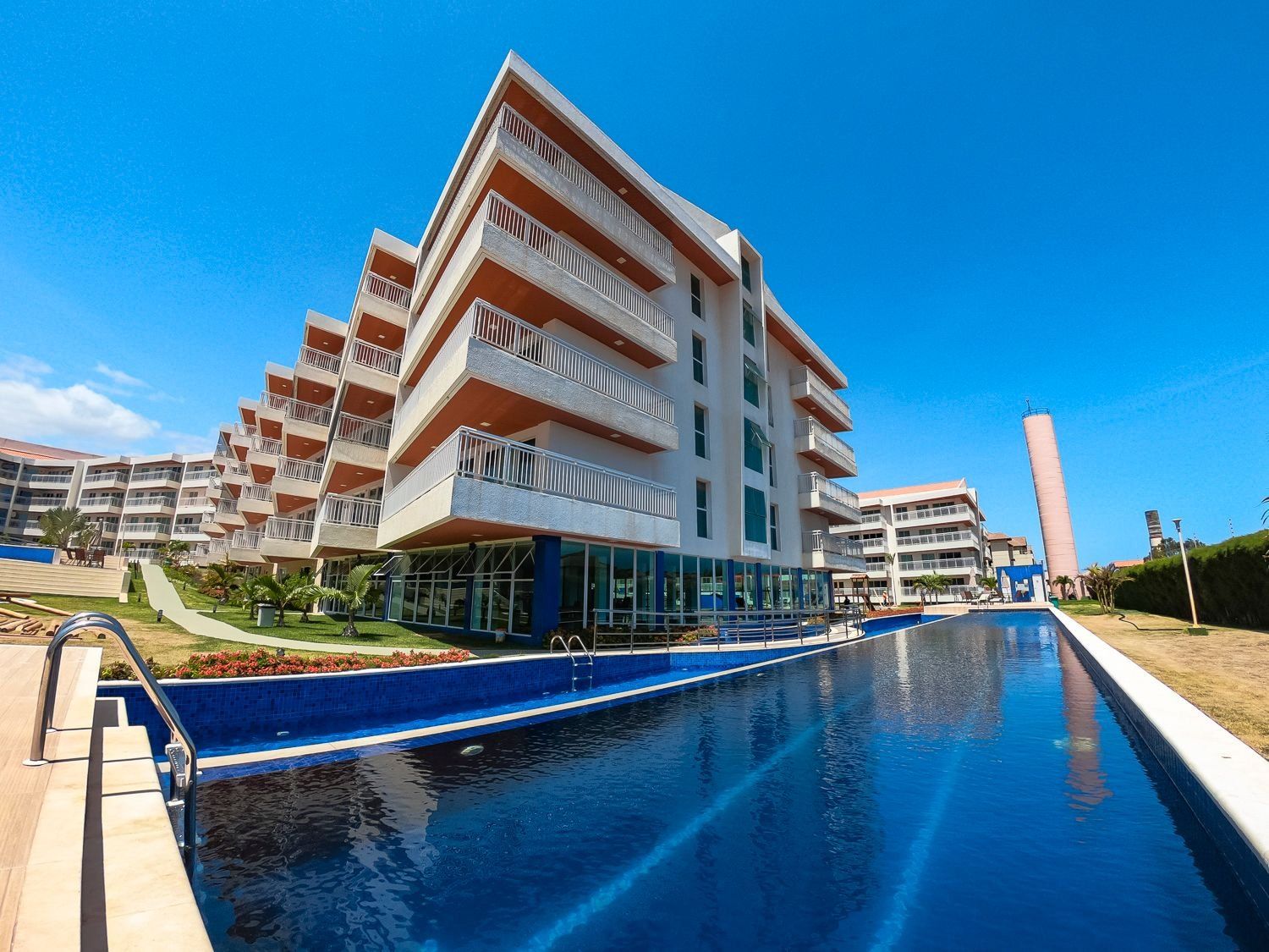 A modern hotel with a pool, multiple balconies, and a clear blue sky.