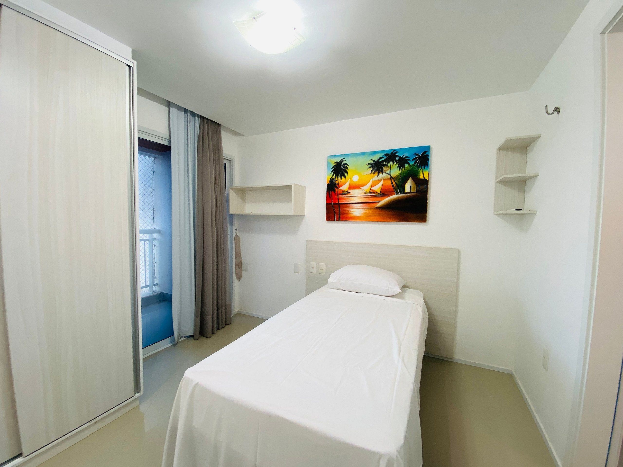 Una habitación de hotel con una cama, un cuadro de un atardecer, una puerta al balcón y una lámpara en el techo.