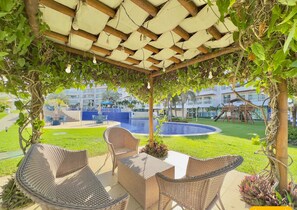 Un espacio de estar al aire libre techado con mobiliario de mimbre, una piscina y vistas a un complejo turístico.