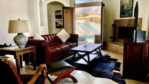 Ein Wohnzimmer mit einem Ledersofa, einem Holztisch, einer Lampe und Blick auf eine Berglandschaft durch ein großes Fenster.