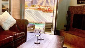 Ein Zimmer mit Blick auf Berge durch ein großes Fenster, eine Ledersofa, ein Holztisch mit zwei Weingläsern und ein Kamin.