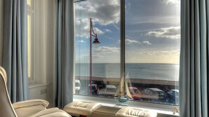 A room with a large window overlooking a parking area and the sea. There are two beige chairs and a small table with a decorative item. - 2 Mareeba (Seaton)