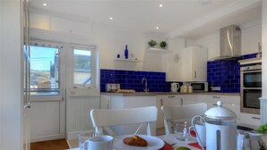 A modern kitchen with white cabinets, blue tiles, and stainless steel appliances. There is a dining table with a plate of food and a coffee pot. - 2 Mareeba (Seaton)