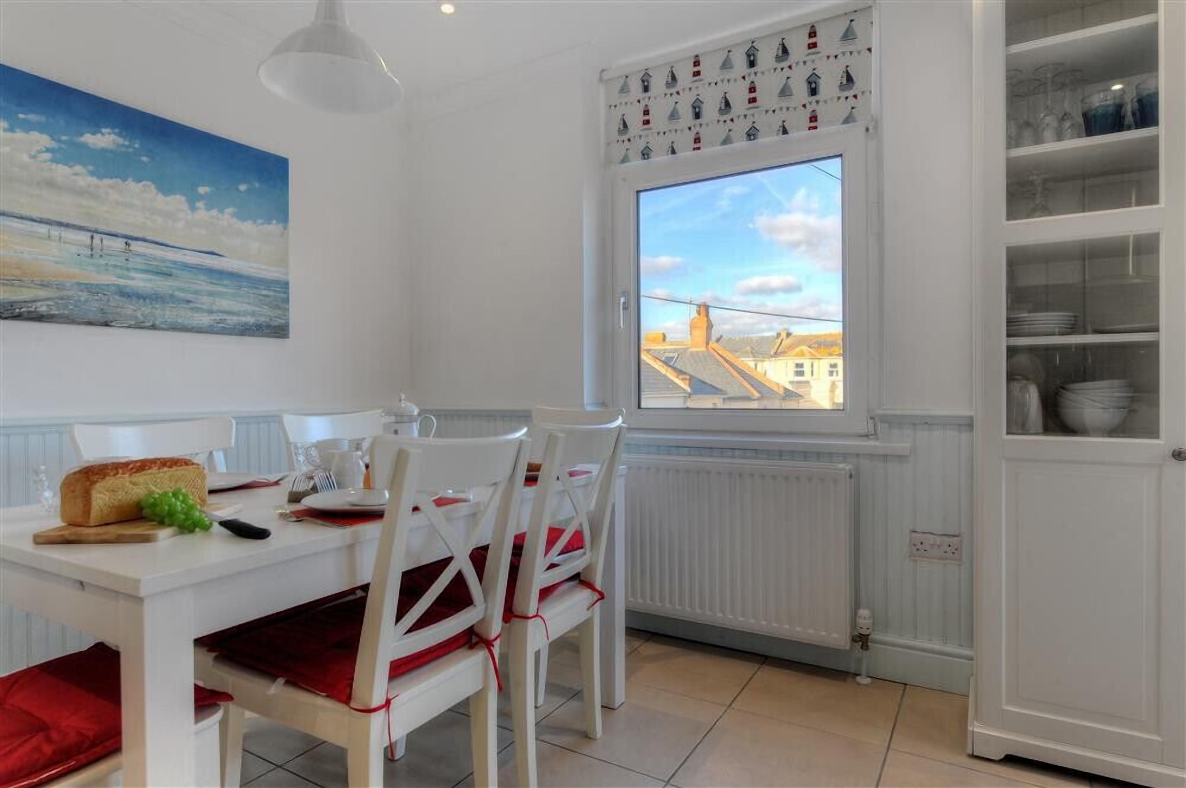 Un comedor con una mesa blanca dispuesta para cuatro, un cuadro de una playa y una ventana con vistas a edificios.