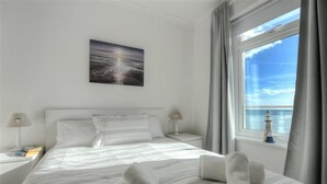 A neatly made bed with a view of the sea through a window, a painting on the wall, and a nightstand with a lamp and books. - 2 Mareeba (Seaton)