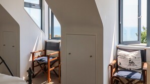 Ein Zimmer mit schrägen Wänden, einem Holzstuhl, einer weißen Tür und großen Fenstern mit Meerblick.