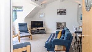 Ein modernes Wohnzimmer mit einem blauen Sofa, einem Holzstuhl, einem Fernseher auf einem Standfuß und einem kleinen Tisch mit einem Hocker.