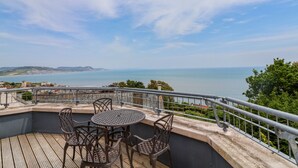 Eine Terrasse mit rundem Tisch und Stühlen, mit Blick auf eine Küstenlandschaft.