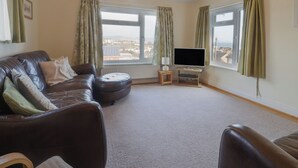 A living room with a leather sofa, a TV on a stand, and large windows with curtains. - Greystones (Lyme Regis)