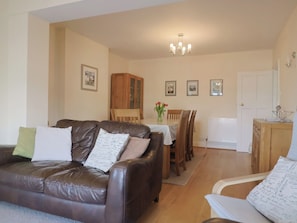 A living room with a brown leather sofa, a dining table with chairs, a wooden cabinet, and a chandelier. - Greystones (Lyme Regis)
