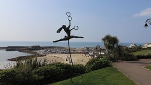 A coastal view with a sculpture of a figure balancing rings, a beach, and a town in the background. - Greystones (Lyme Regis)