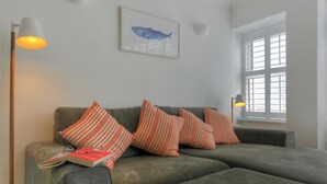 Ein modernes Wohnzimmer mit einem grauen Ecksofa, gestreiften Kissen, einem Bild mit Fischmotiv, einer Stehlampe und einem Fenster mit Jalousien.