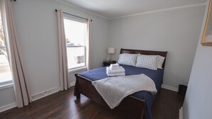 Een opgemaakt bed met een blauwe sprei en witte kussens, een houten hoofdbord, een nachtkastje met een lamp en een raam met gordijnen.