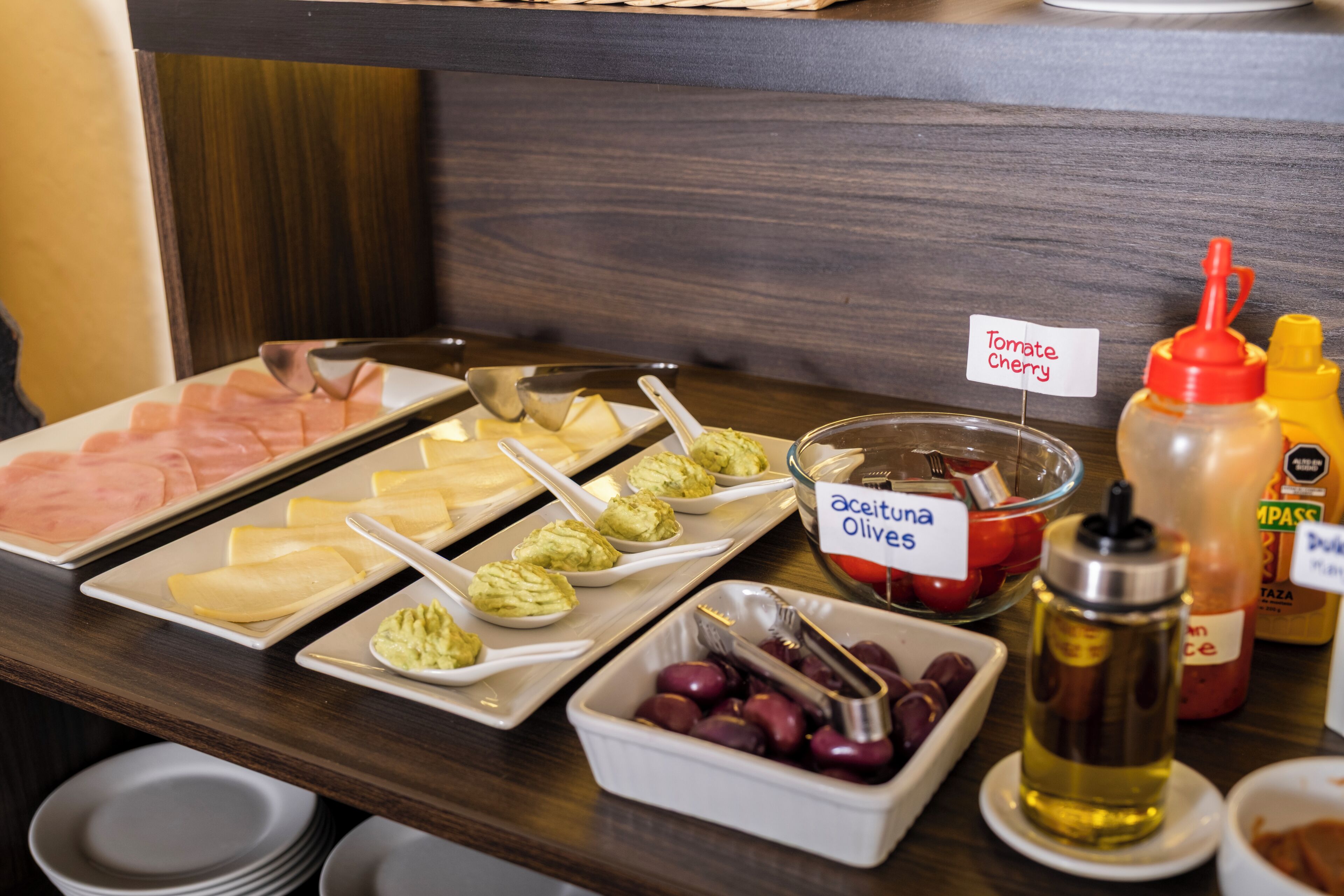 Un bufé con diversas opciones de comida, incluyendo jamón, queso, aceitunas y un bol con la etiqueta 'aceituna Olives'.