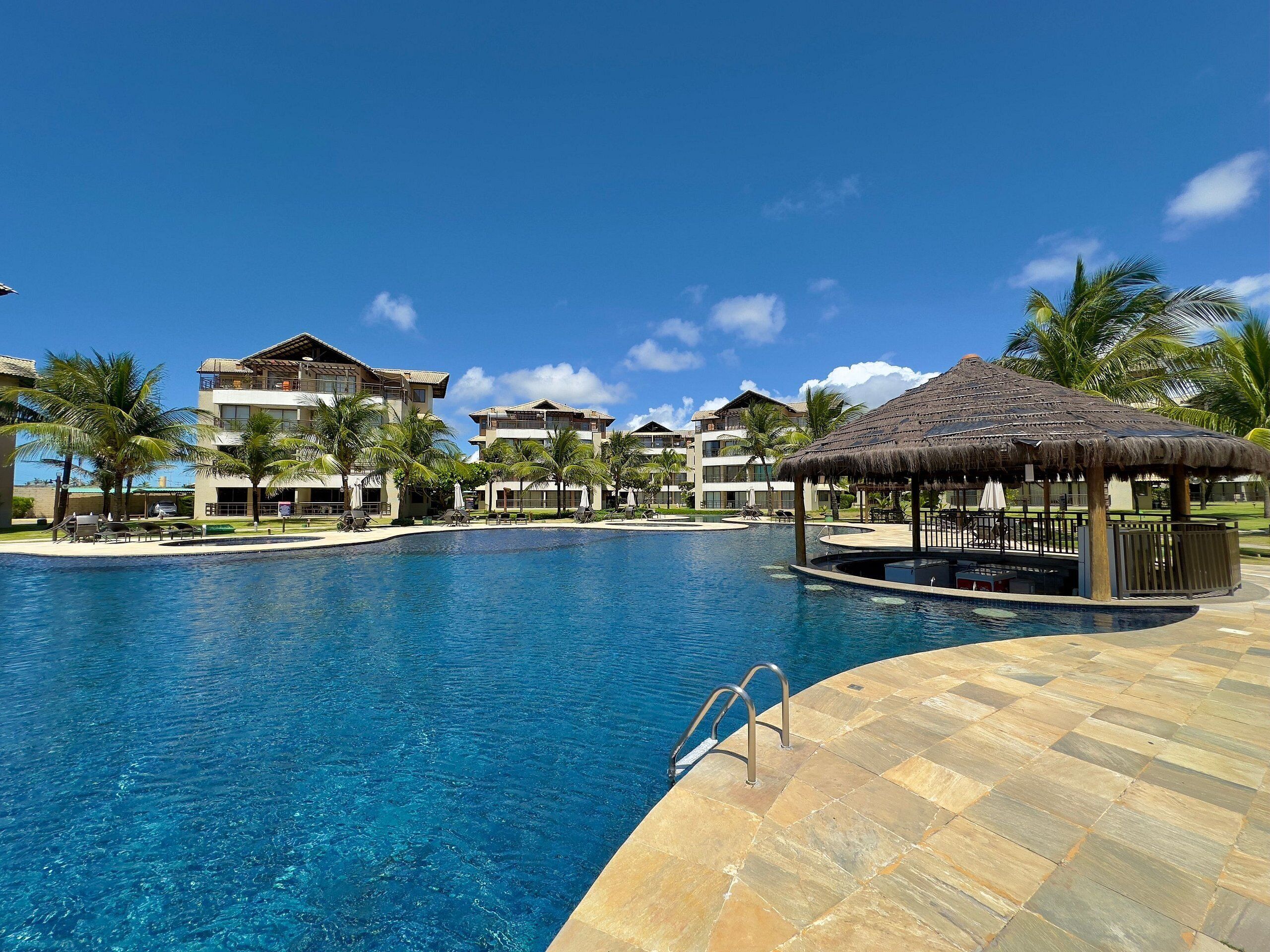 A resort pool with a thatched gazebo, palm trees, and modern buildings in the background.