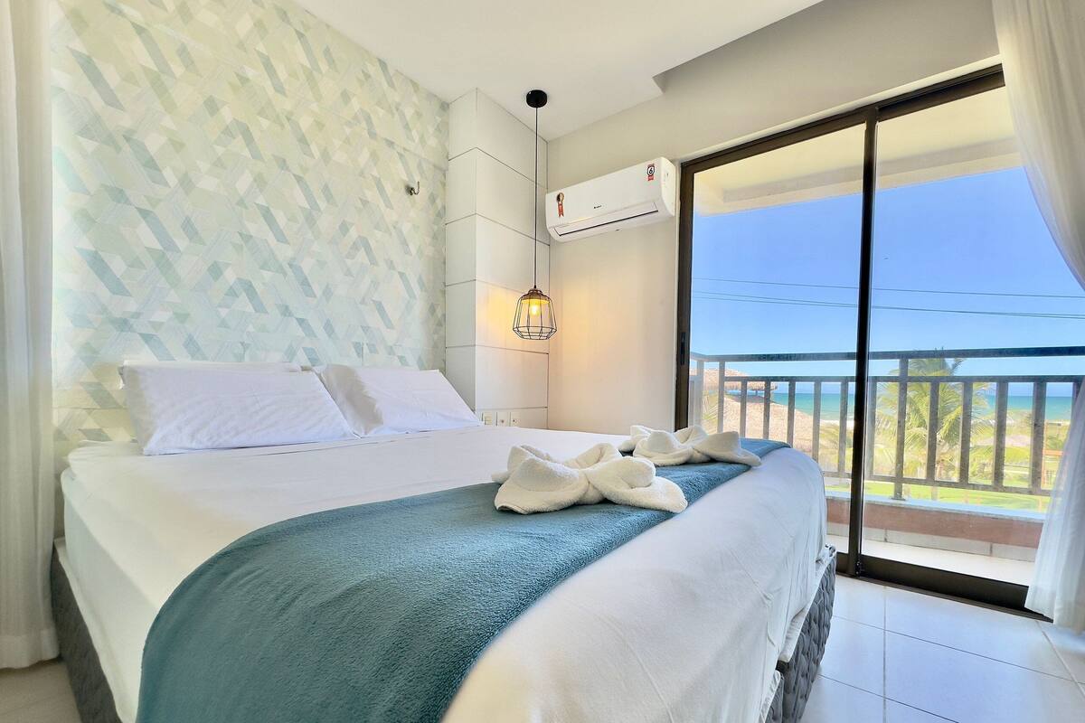 A hotel room with a bed, a view of the beach, and an air conditioning unit.