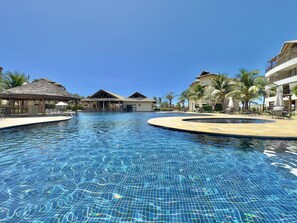 Una piscina con superficie de agua azul cristalina, rodeada de sillones de descanso y sombrillas, con edificios y palmeras al fondo.