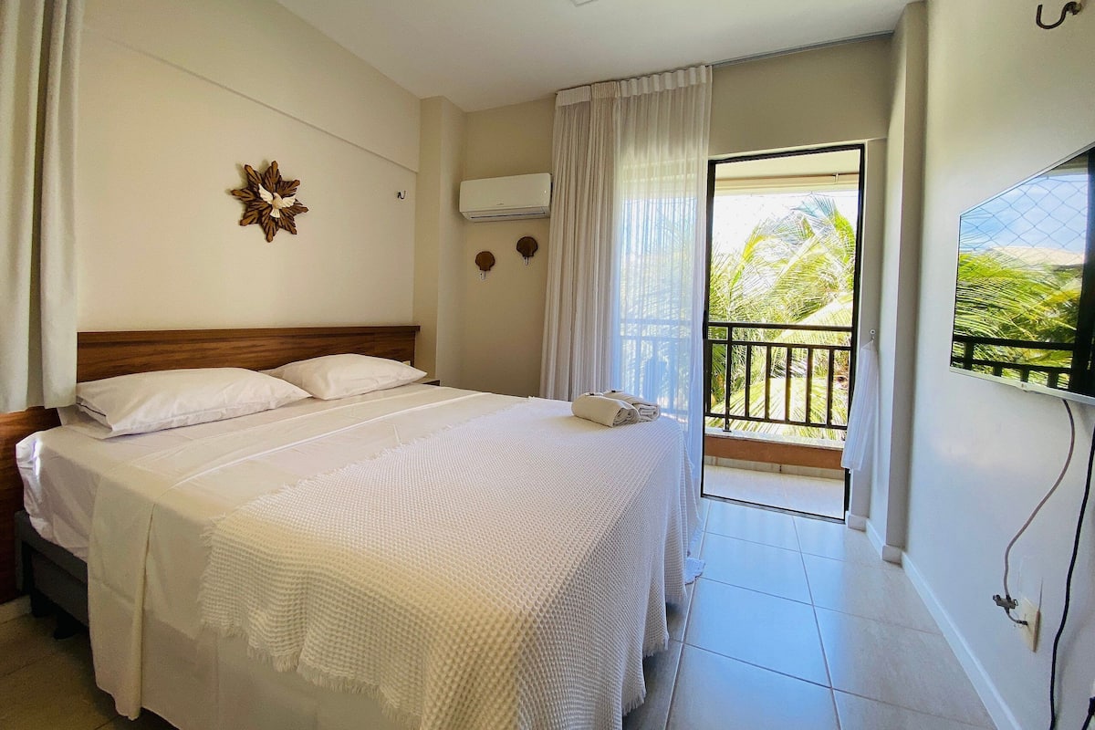 A hotel room with a bed, a window with curtains, a wall-mounted air conditioner, and a balcony with a view of palm trees.