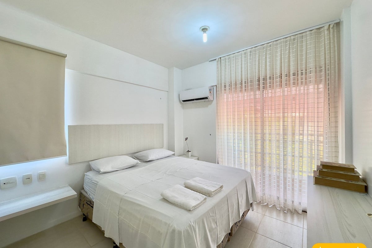 A bedroom with a bed, bedside tables, an air conditioning unit, and a window with curtains.