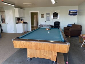 A pool table in a room with a kitchen area, a dartboard, and a couch.