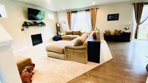A living room with a beige sectional sofa, a flat-screen TV mounted on the wall, a fireplace, and a wooden floor.