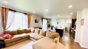 A modern living room with a large beige sectional sofa, a dining area with a kitchen in the background, and a view of the outdoors through large windows.
