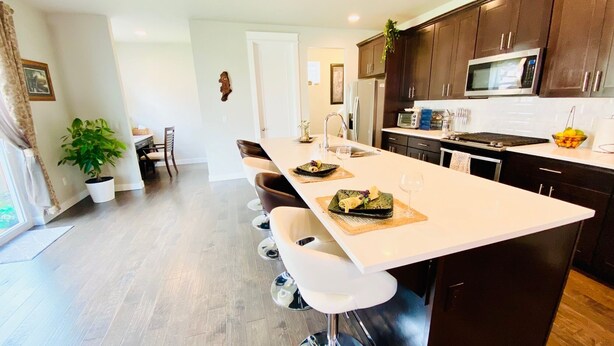 A modern kitchen with dark wooden cabinets, a white countertop, and stainless steel appliances. There is a dining area with a long table and white chairs.