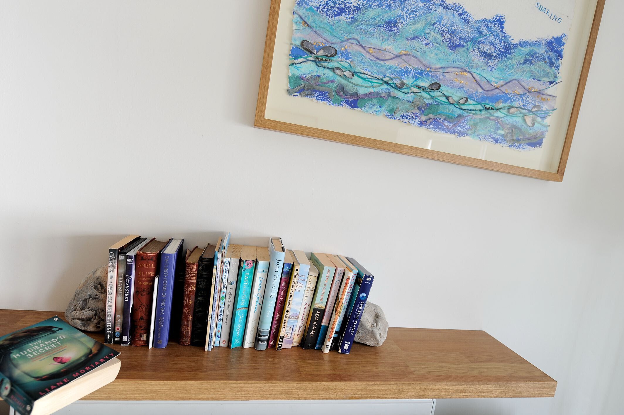 Une bibliothèque remplie de divers ouvrages et, au mur, un tableau abstrait encadré.