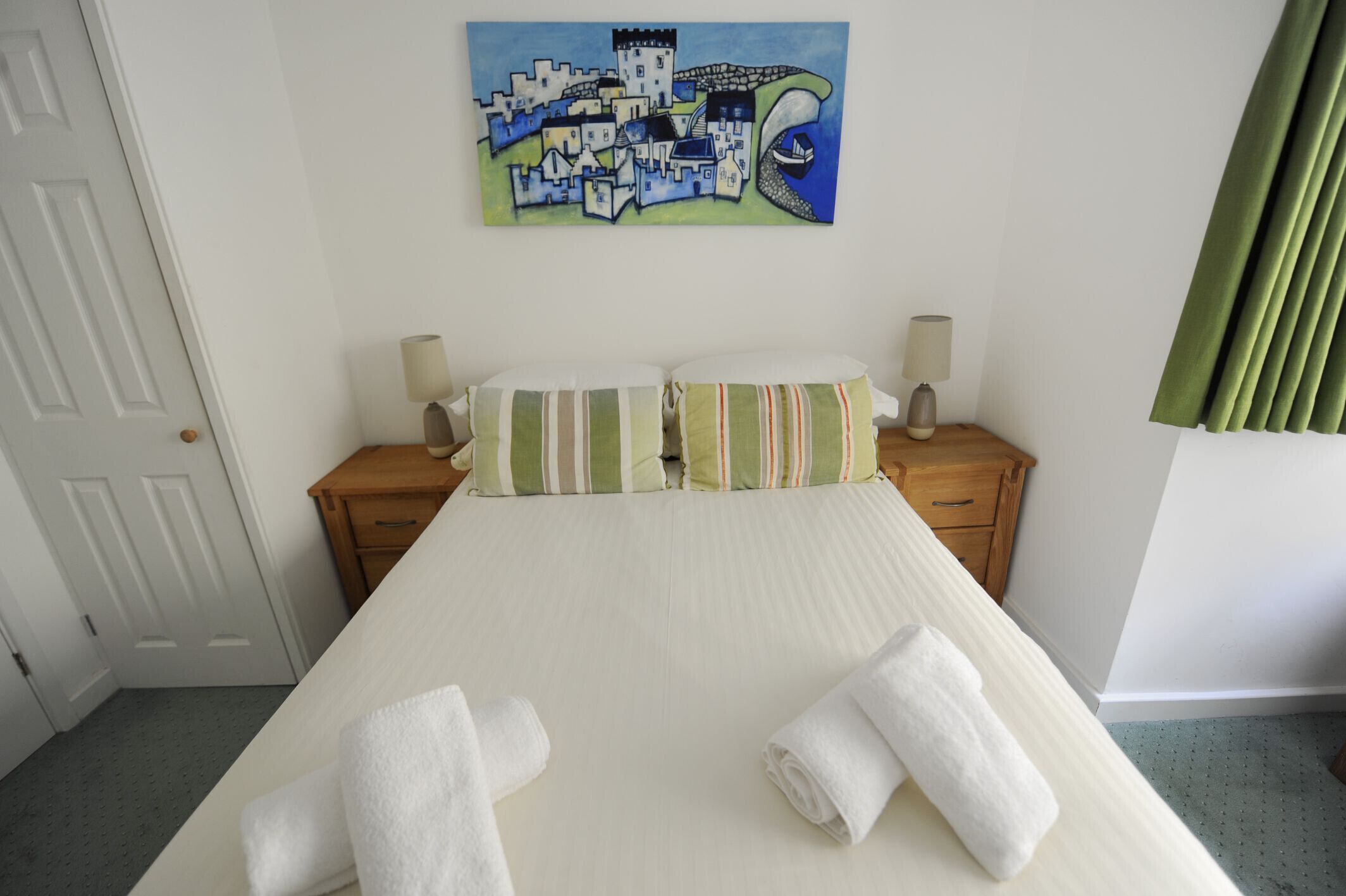 A neatly made bed with striped pillows, a painting of a coastal town on the wall, and a wooden nightstand with a lamp.