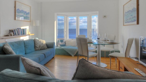 Una sala de estar con un sofá azul, una mesa de comedor de vidrio y vista al mar a través de grandes ventanales.