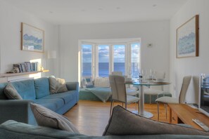 A living room with a blue sofa, a glass dining table, and a view of the sea through large windows. - Sea Fever (Lyme Regis)