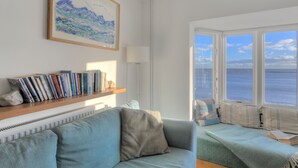 Un salón con un sofá, una estantería llena de libros, una lámpara y un ventanal con vistas al mar.