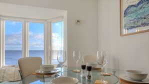 Comedor con una mesa de vidrio, platos y tazones blancos, y copas de vino. Se aprecia vista al mar a través de amplias ventanas.