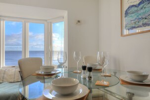 Comedor con una mesa de vidrio, platos y tazones blancos, y copas de vino. Se aprecia vista al mar a través de amplias ventanas.