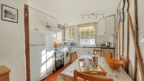 A kitchen with white cabinets, a stove, a microwave, and a sink. There is a dining table with chairs and a window with blinds. - Waltham House (Lyme Regis)