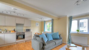 A compact living space with a kitchenette, a grey sofa with blue cushions, a wooden dining table, and a window with curtains. - The Old Bakehouse (Lyme Regis)