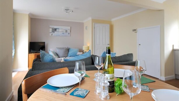 A dining table set with plates, glasses, and a bottle of wine, with a sofa and television in the background. - The Old Bakehouse (Lyme Regis)