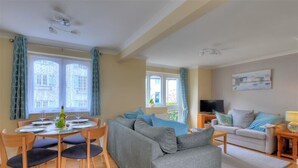 A living room with a grey sofa, a dining table with chairs, and a TV on a stand. - The Old Bakehouse (Lyme Regis)