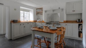A kitchen with white cabinetry, a wooden table, and chairs. There is a window providing natural light, and a stove is visible in the background. - 12 Mill Green (Lyme Regis)