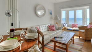Un salon avec un canapé blanc, des fauteuils en osier, une table basse en bois et une vue sur la mer grâce à de grandes fenêtres.