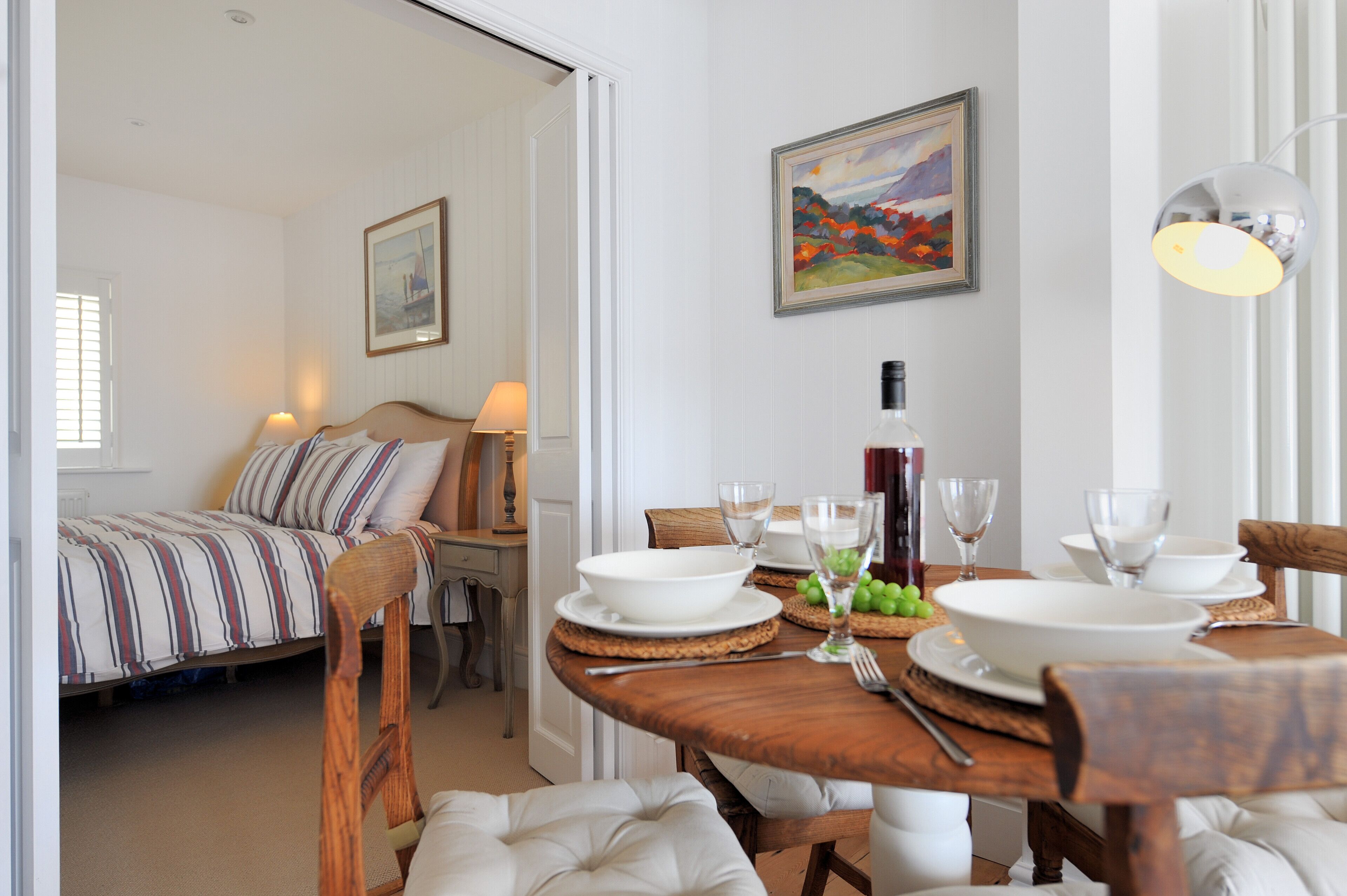 A dining area with a wooden table set for two, a bed with striped bedding, and a painting on the wall.